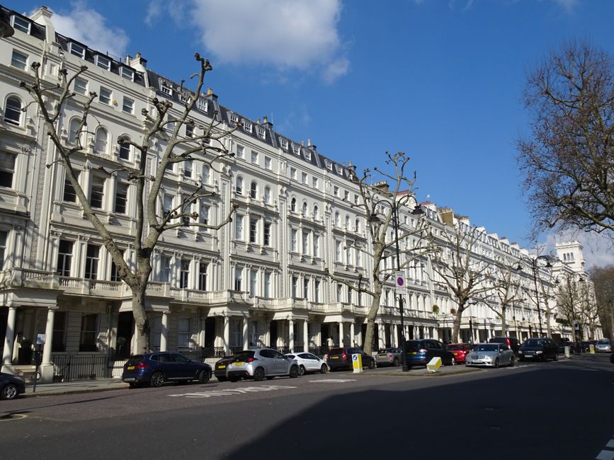 A row of elegant Victorian-style terraced buildings with white facades and intricate architectural details, situated along a tree-lined street in Maida Vale. The trees are leafless, indicating a winter or early spring setting, with their bare branches extending upward. Cars of various colors are parked along the curb, some with visible license plates, under a bright blue sky with a few scattered clouds. The scene captures a quiet urban residential area, emphasizing clean surfaces and well-maintained exteriors, reflecting the local environment where professional domestic cleaning services, such as those provided by Maida Vale Carpet Cleaning, may operate.