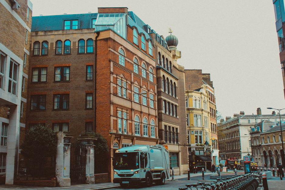 A city street in Maida Vale featuring a mix of historic and modern buildings, including a red brick structure and a cream-colored building with ornate architectural details. The street is lined with cars and bicycles, and a blue maintenance vehicle is parked on the side. The scene is lit by natural daylight, highlighting the clean, well-maintained surfaces of the buildings and pavement, with some reflections visible on the glass windows. The area appears tidy with no visible litter or dust, consistent with residential and commercial surface cleaning and deep cleaning standards that Maida Vale Carpet Cleaning offers, especially relevant for addressing delays and access issues affecting local cleaning services.