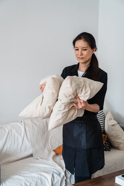 A woman with dark hair, dressed in a black uniform, holds two large beige pillows with clean, fabric covers in a well-lit living room. The room features a white sofa with a cream-colored cushion and a patterned throw pillow, a wooden side table, and a plain light-colored wall background. The lighting highlights the surfaces' cleanliness and the neat arrangement of the cushions. This image pertains to domestic surface cleaning, highlighting professional upholstery and fabric maintenance, as seen in the Clifton Villas upholstery cleaning before and after case study by Maida Vale Carpet Cleaning.