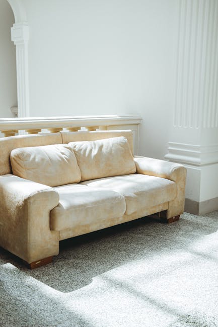 A beige fabric sofa with plush cushions positioned against a white wall in a well-lit living room. The corner of the sofa shows clean, smooth upholstery indicating recent deep cleaning or surface sanitisation by Maida Vale Carpet Cleaning. The carpet beneath the sofa is light grey with a soft texture, appearing freshly vacuumed and free of dust or stains. Natural light from an unseen window casts soft shadows on the carpet, highlighting the tidy and maintained condition of the space. Visible architectural details include white painted panel moulding and a decorative column base, emphasizing a clean and classic interior setting. This image exemplifies professional domestic cleaning and surface maintenance to ensure a hygienic, inviting environment, as showcased in the Clifton Villas upholstery cleaning before and after case study on maidavalecarpetcleaning.org.uk.