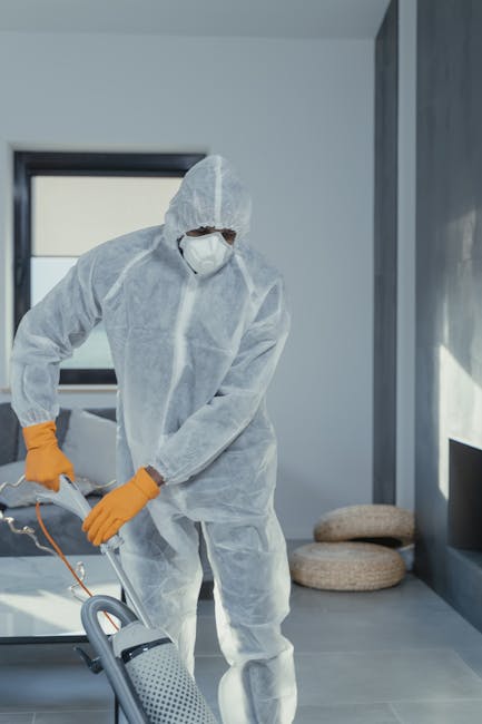 A professional cleaner dressed in a white protective suit, face mask, and orange gloves is performing surface cleaning in a modern, well-lit living room. The room features light-colored flooring, possibly ceramic or tiles, and has large windows allowing natural light to fill the space. In the background, a pair of beige cushions are placed on the floor near a dark-colored wall. The cleaner is using a vacuum or cleaning device to maintain hygiene and cleanliness, demonstrating a deep cleaning process suitable for residential or commercial environments, reflecting the expertise of Maida Vale Carpet Cleaning in emergency carpet stain removal and surface sanitisation. The overall scene emphasizes thorough cleanliness and maintenance of a tidy, hygienic living space.