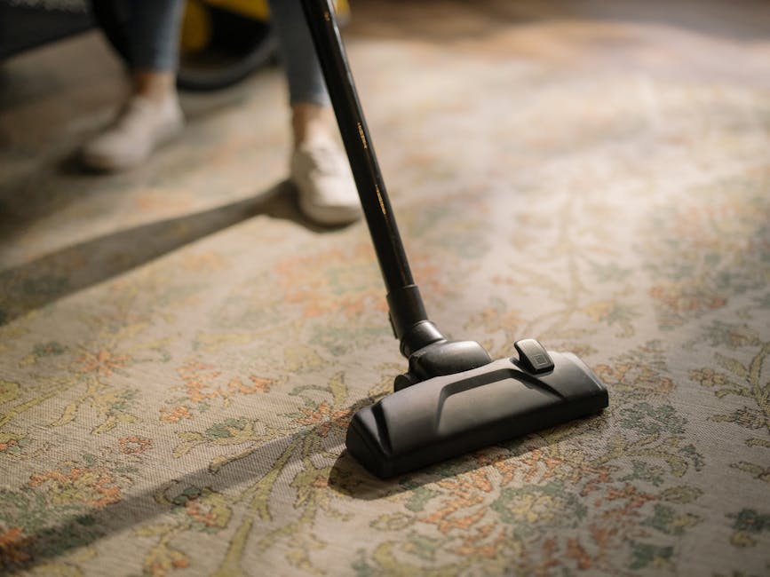 A professional cleaner dressed in a white protective suit, face mask, and orange gloves is performing surface cleaning in a modern, well-lit living room. The room features light-colored flooring, possibly ceramic or tiles, and has large windows allowing natural light to fill the space. In the background, a pair of beige cushions are placed on the floor near a dark-colored wall. The cleaner is using a vacuum or cleaning device to maintain hygiene and cleanliness, demonstrating a deep cleaning process suitable for residential or commercial environments, reflecting the expertise of Maida Vale Carpet Cleaning in emergency carpet stain removal and surface sanitisation. The overall scene emphasizes thorough cleanliness and maintenance of a tidy, hygienic living space.