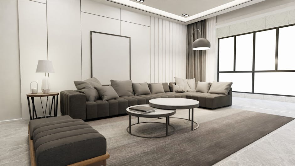 A spacious, modern living room featuring a large L-shaped gray sofa with multiple cushions, positioned on a neutral-toned area rug. In front of the sofa are two round nested coffee tables, one holding a closed book. To the left, a small wooden side table with a white lamp sits beside the sofa. The room has sleek white walls, a minimalistic white ceiling with built-in lighting, and expansive black-framed windows allowing natural light to illuminate the space. The floor appears to be a light-colored tile or stone, contributing to the room's clean and bright aesthetic. This setting exemplifies thorough surface cleaning and maintenance, emphasizing clarity and hygiene, and is consistent with professional domestic cleaning standards provided by Maida Vale Carpet Cleaning for effective sanitisation and deep cleaning of living environments.