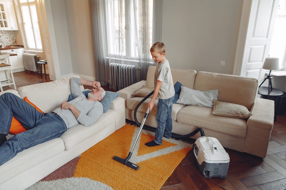 A young boy using a vacuum cleaner to deep clean a beige fabric sofa in a living room with large windows, sheer curtains, and a grey wall. The vacuum cleaner is connected to a small, portable machine on the wooden floor, which is partially covered by a multicolored rug. An elderly man with grey hair and a beard, dressed in a grey sweater and blue jeans, is lying on the sofa, resting with his eyes closed. The room features natural light, a small table with a lamp beside the sofa, and additional seating in the background, reflecting a clean and well-maintained domestic setting with a focus on surface cleaning and hygiene, as highlighted in the guide by Maida Vale Carpet Cleaning.