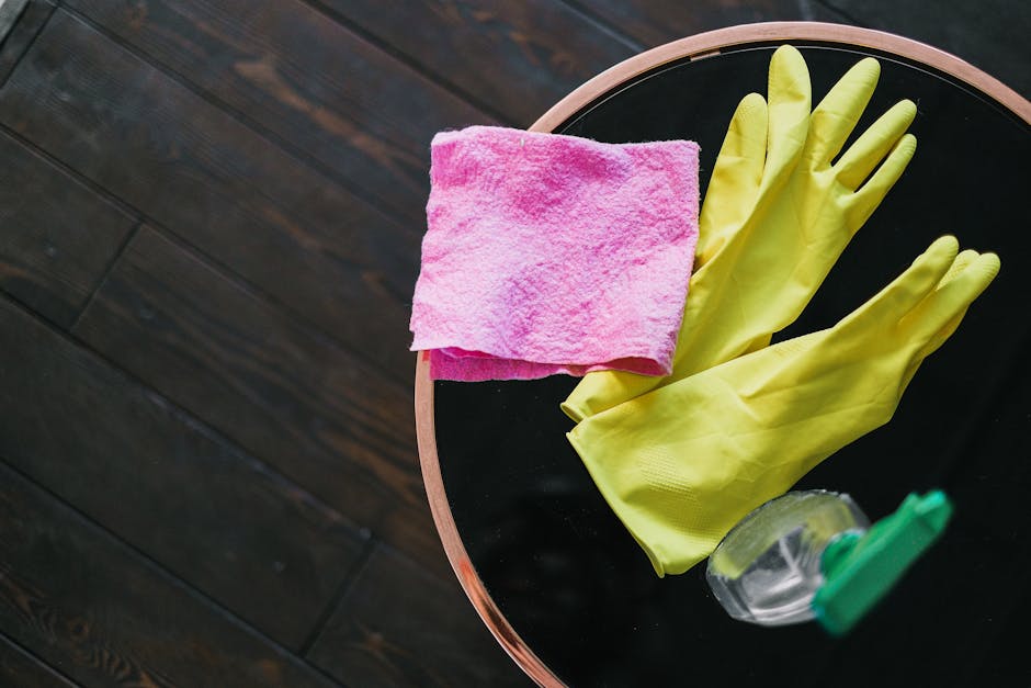 A round black table with a light wooden edge in a room with polished dark wooden flooring. On the table, there is a pair of bright yellow rubber cleaning gloves, a folded pink microfiber cloth, and a clear spray bottle with a green trigger, all arranged for cleaning purposes. The surface of the table appears clean and free of dust or stains, indicating readiness for surface cleaning or sanitisation tasks, consistent with professional domestic cleaning standards. The lighting in the room highlights the reflective surface of the table and the vibrant colors of the cleaning tools, emphasizing a well-maintained environment. This setup is representative of cleaning equipment used by Maida Vale Carpet Cleaning for thorough hygiene and maintenance, aligning with landlord cleaning obligations and tenancy rules in Maida Vale.