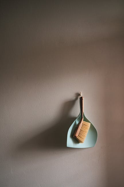 A light blue silicone wall hook mounted on a plain, neutral-toned wall. Hanging from the hook are two cleaning tools: a bamboo-handled scrub brush with stiff bristles and a soft brown cloth. The wall surface appears smooth and clean, with natural lighting casting subtle shadows that highlight the textures of the brush and cloth. This setup suggests a space designated for surface cleaning or general household tidying, typical of domestic cleaning routines. The arrangement is minimalistic, emphasizing the importance of proper storage for cleaning equipment. The presence of the Maida Vale Carpet Cleaning company's branding indicates the context of professional domestic or commercial cleaning services associated with the page about landlord cleaning obligations and tenancy rules for Maida Vale residents.