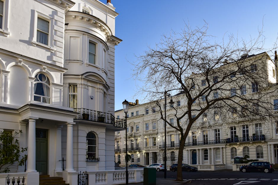 Exterior view of a row of elegant white residential buildings with ornate detailing, large sash windows, and decorative balconies. The buildings feature classic architectural elements including pilasters, curved bay windows, and a rounded corner turret. In the foreground, a leafless tree with numerous branches stands beside a traditional black street lamp. Several parked cars are visible along the street, which is lined with a black metal fence. The scene is illuminated by bright daylight, highlighting the clean, well-maintained facades and the clear blue sky above. This setting exemplifies the pristine condition of Victorian-style residential properties, suitable for professional surface cleaning or deep cleaning services by Maida Vale Carpet Cleaning.