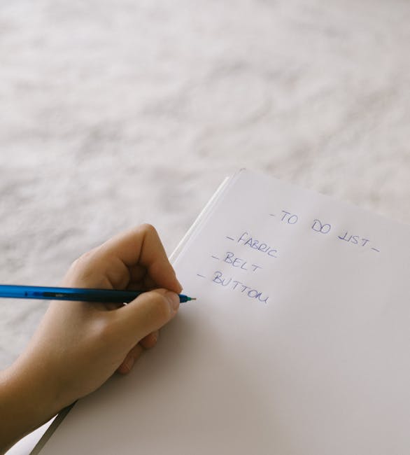 A person's left hand is holding a blue pen and writing on a white notepad, creating a checklist with items including 'fabric,' 'belt,' and 'button.' The notepad is resting on a light-colored surface, possibly a table or countertop, with no other objects visible in the frame. The scene is well-lit with natural or bright artificial lighting, emphasizing the clarity and neatness of the handwritten list, which appears to be related to preparing for cleaning or organising tasks. This image visually conveys the process of planning or note-taking for domestic or commercial cleaning services provided by Maida Vale Carpet Cleaning, in line with their spring cleaning checklist for 2027 featured on their website.