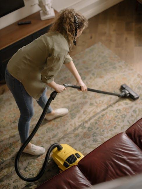 A woman with curly hair wearing a grey jacket, dark jeans, and white sneakers is performing surface cleaning in a living room. She is using a yellow vacuum cleaner with a long, black hose and a cleaning head attachment to deep clean a patterned, beige and blue area rug placed on a wooden floor. The room features a wooden sideboard along the wall with decorative items, and natural light illuminates the space, highlighting the carpet's cleanliness. The scene depicts domestic cleaning, emphasizing hygiene and maintenance by Maida Vale Carpet Cleaning, which specializes in carpet and surface sanitisation services in Maida Vale.