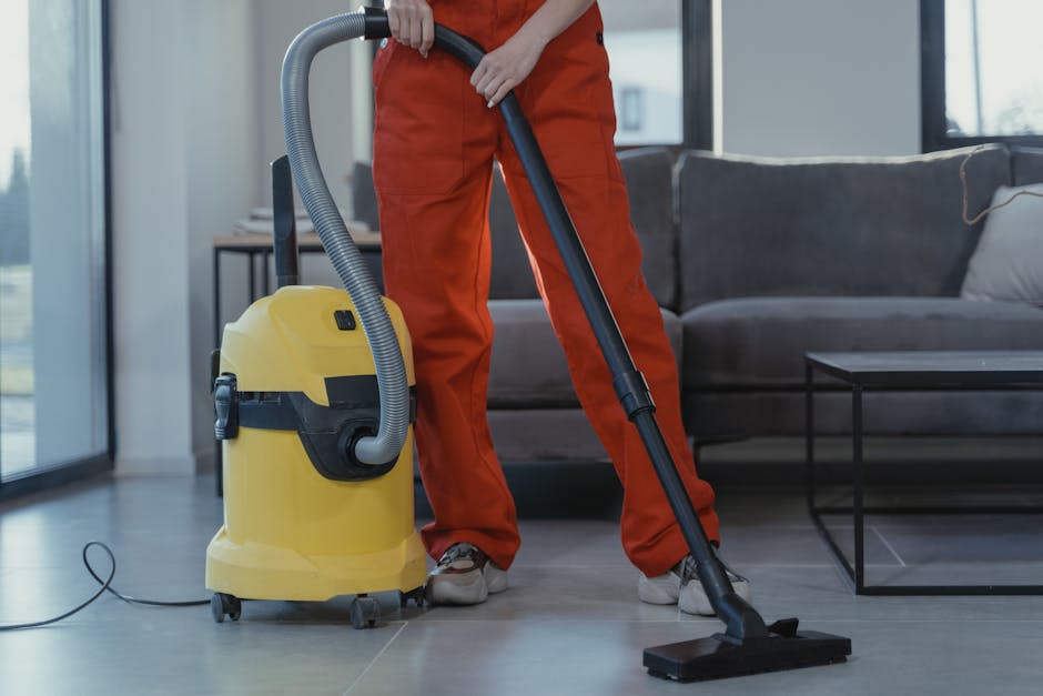 A person wearing orange overalls is operating a yellow commercial vacuum cleaner with a flexible grey hose, performing surface cleaning on a tiled floor in a modern living room. The room features a dark grey sofa with cushions, a black metal coffee table, and large windows allowing natural light to brighten the space. The floor appears clean and dry, with the vacuum nozzle pressed flat against the surface, indicating active deep cleaning or sanitisation. Visible objects include the vacuum unit on wheels positioned near the person's feet, and the room's decor emphasizes a tidy, well-maintained environment as part of professional domestic cleaning services by Maida Vale Carpet Cleaning.
