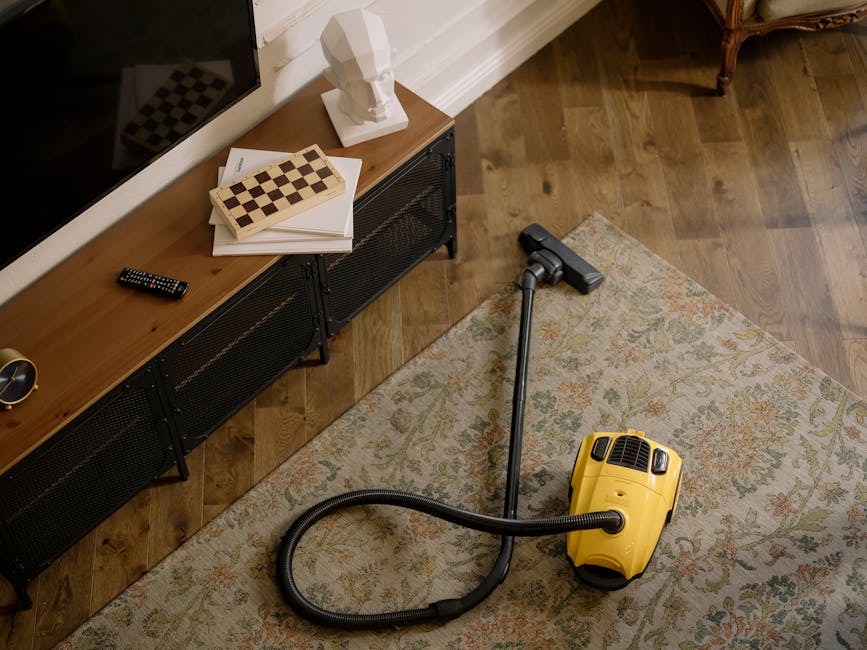 Interior scene showing a yellow canister vacuum cleaner with a flexible hose and cleaning head on a patterned area rug, situated on a wooden floor in a living room. To the left, a low black media unit holds a remote control, a white bust sculpture, and a stack of papers and chessboard. The room is well-lit with natural light, and the overall appearance is clean and well-maintained, reflecting professional surface cleaning and deep cleaning practices provided by Maida Vale Carpet Cleaning for period home carpets on Sutherland Avenue.