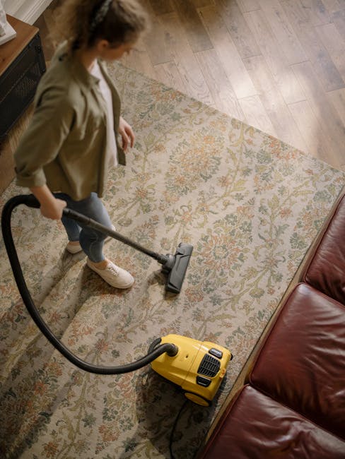 A woman vacuuming a patterned area rug in a living room, demonstrating surface cleaning as part of domestic carpet maintenance. The room features wooden flooring surrounding the rug, a red leather sofa on one side, and natural lighting illuminating the space. The vacuum cleaner is yellow with a black hose, and the woman is dressed casually in a beige jacket, jeans, and sneakers. The rug displays a floral design in muted tones, and the scene highlights the thorough cleaning process carried out by Maida Vale Carpet Cleaning, specializing in deep cleaning and sanitisation of carpets in period homes along Sutherland Avenue.