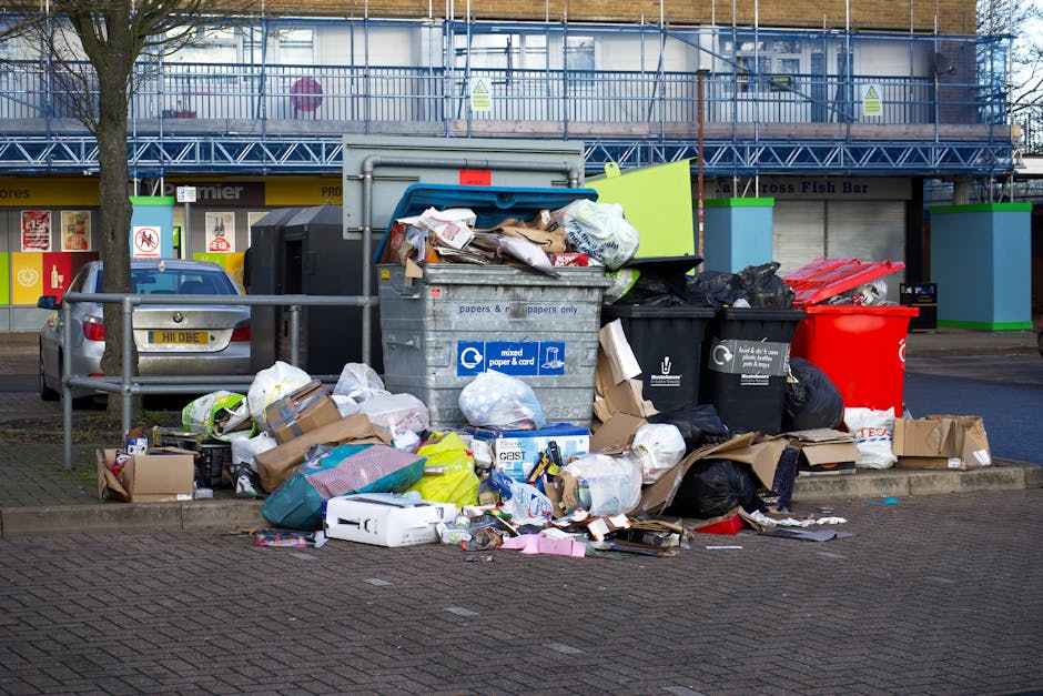 An outdoor scene showing a large collection of discarded household waste and recycling bins on a paved sidewalk in front of a commercial building with a blue metal scaffolding structure. The waste includes mixed paper and cardboard boxes, black and red plastic trash bags, flattened cardboard packaging, and loose paper items, some spilling onto the ground. The areas are cluttered and unkempt, indicating improper disposal of rubbish. Nearby, a parked silver car is partially visible, with a metal railing separating the sidewalk from the street. Bright daytime lighting illuminates the scene, emphasizing the scattered waste. This image highlights the importance of proper waste management and cleaning in urban environments, in line with Maida Vale Carpet Cleaning’s emphasis on surface cleaning, hygiene, and environmental maintenance, especially relevant to Westminster Council rules for mattress disposal in Maida Vale.