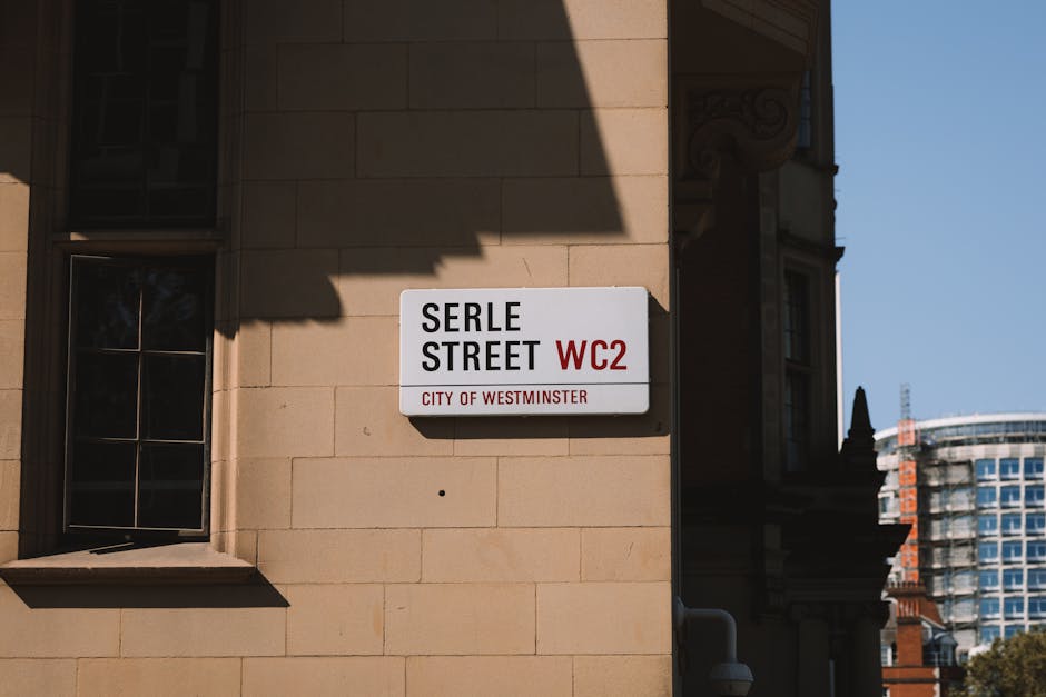 Close-up view of a street sign on a beige stone building wall displaying 'Serle Street WC2' in black and red lettering, with a small window with dark frames visible on the left, and a shadow cast over part of the sign. In the background, buildings with modern glass facades and architectural details under a clear blue sky are visible. The scene is outdoors, illuminated by natural daylight, and the area appears clean and well-maintained, consistent with standard urban street characteristics. Maida Vale Carpet Cleaning emphasizes surface cleaning and hygiene in their residential and commercial cleaning services, aligning with regulations like Westminster Council's rules for mattress disposal.